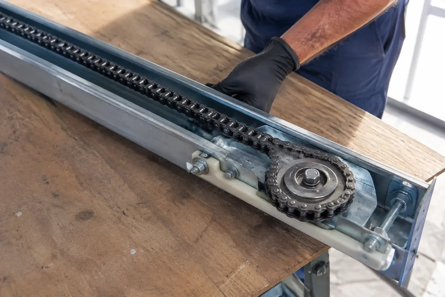 Gloved hand resting on a steel opener rail laid on a wooden bench, showing a drive chain looped around a sprocket wheel. — Chain-Drive Opener Repair, St. Clements